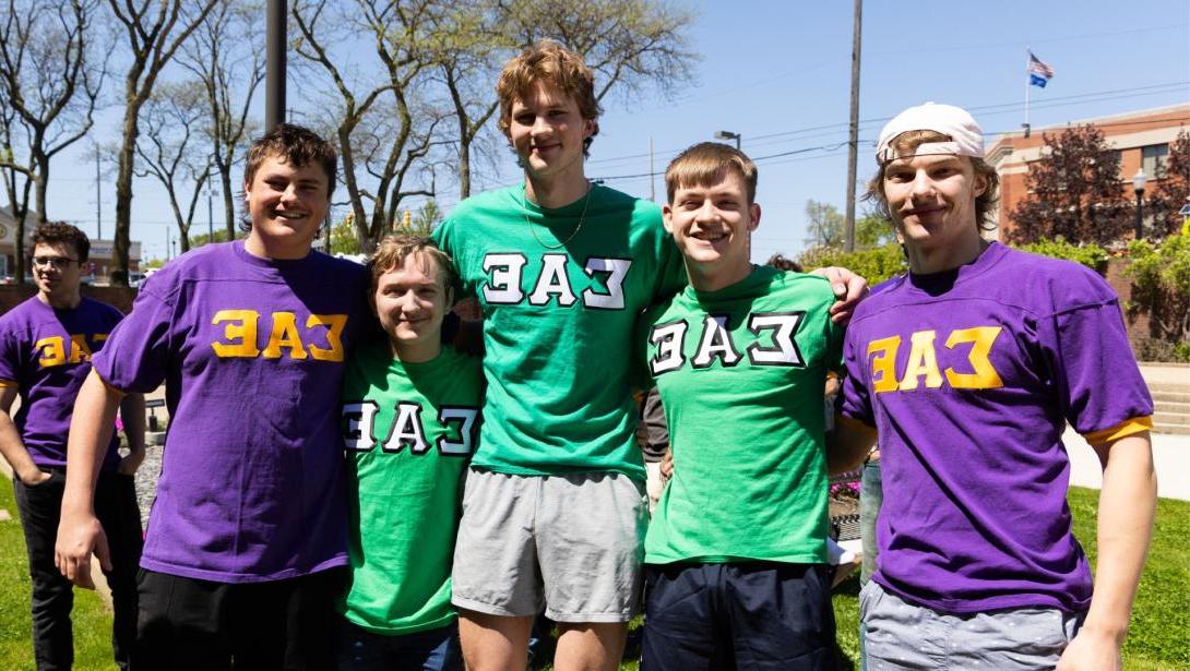 Five Kettering students standing outside smiling while wearing purple and green Sigma Alpha Epsilon fraternity shirts